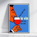 A framed art print leaning against a white wall on a light wood floor. The print features a retro-style illustration of a man with an orange face holding a glass of red wine against a blue background, with the French text 'qui aime le vin aime la vie'.