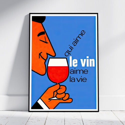 A framed art print leaning against a white wall on a light wood floor. The print features a retro-style illustration of a man with an orange face holding a glass of red wine against a blue background, with the French text 'qui aime le vin aime la vie'.