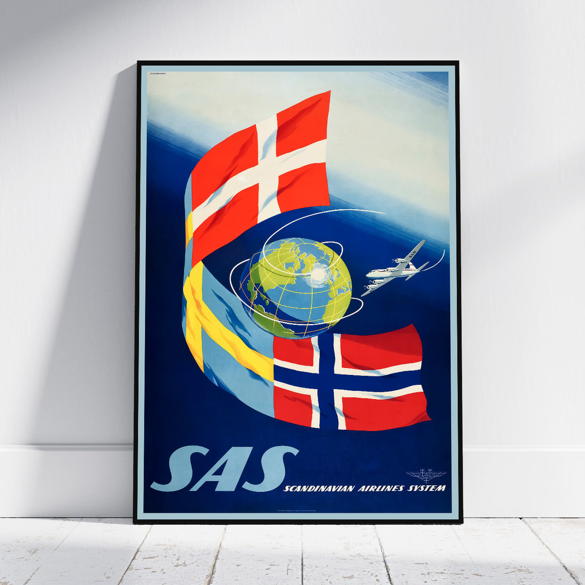 A framed vintage-style poster leaning against a white wall on a light wood floor. The poster features the text 'SAS' and 'SCANDINAVIAN AIRLINES SYSTEM' with large red-and-white Scandinavian flags, a blue globe, and a small airplane illustration.
