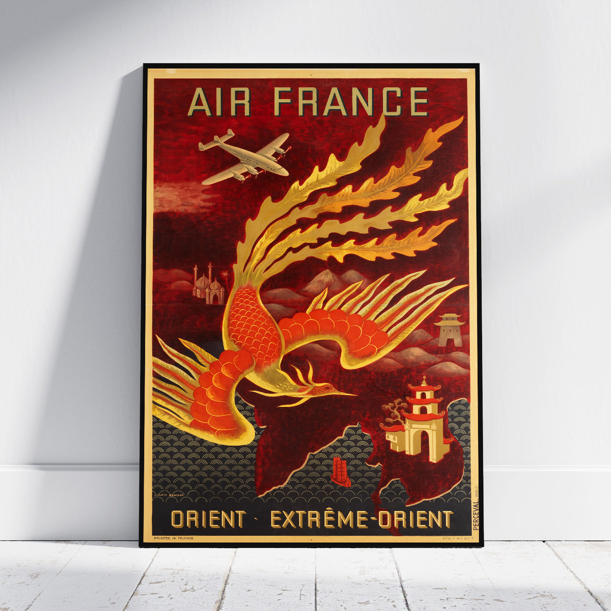 A vintage-style Air France travel poster leaning against a white wall on a light wood floor. The poster features a large red and gold phoenix, an airplane, and the text 'AIR FRANCE' at the top and 'ORIENT EXTRÈME-ORIENT' at the bottom.