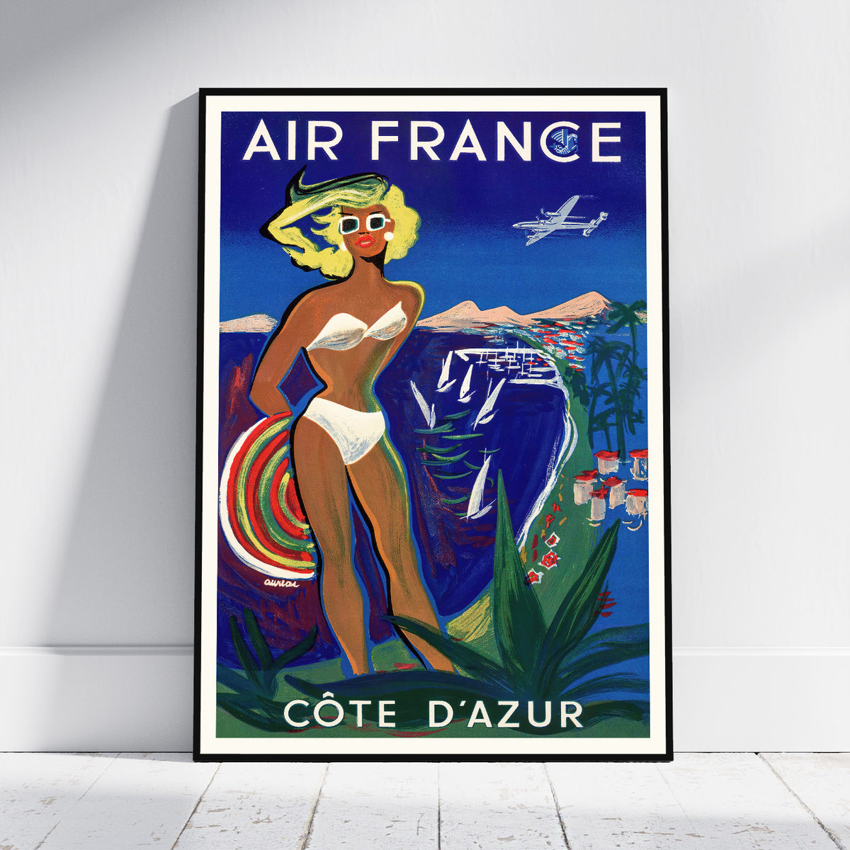 A framed vintage-style Air France travel poster leaning against a white wall on a light floor. The poster features the text 'AIR FRANCE' at the top and 'CÔTE D'AZUR' at the bottom, with an illustrated woman in a white bikini holding a striped surfboard, a blue airplane, and coastal scenery.