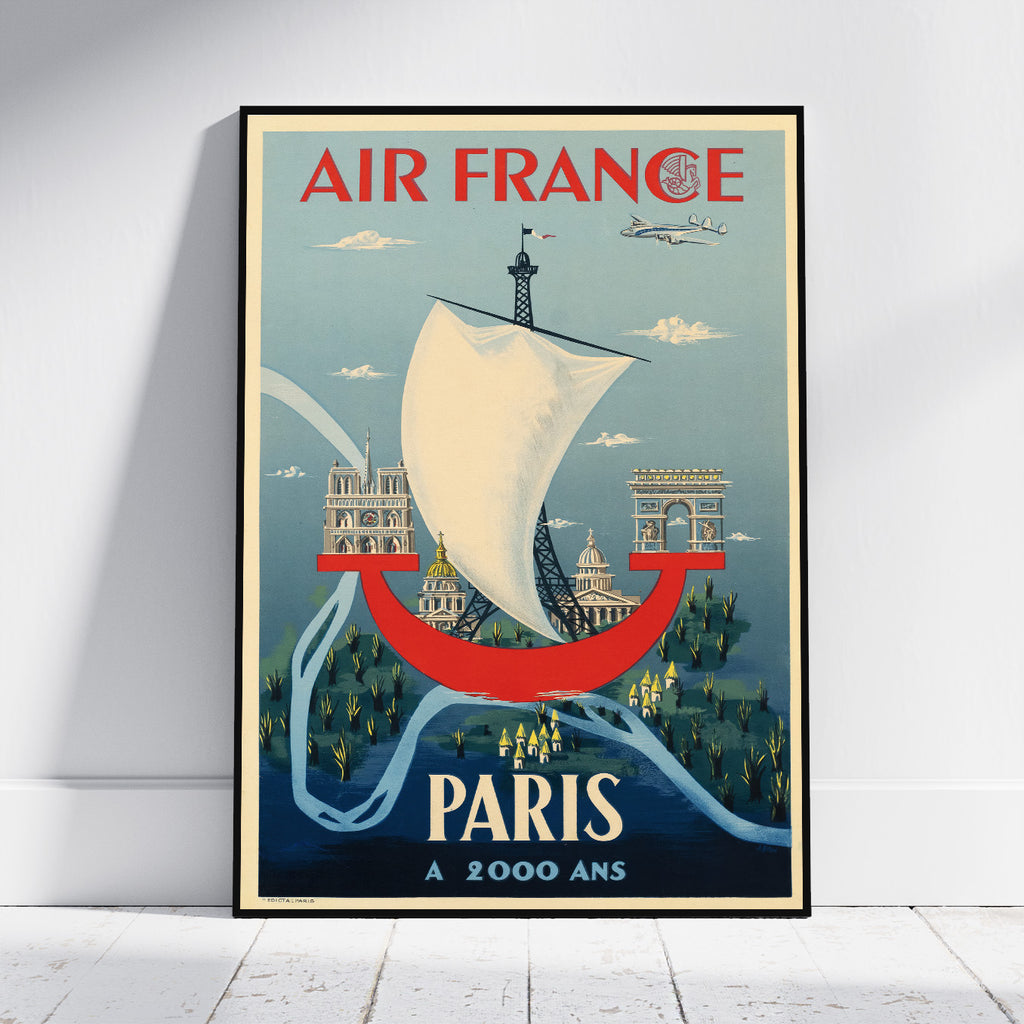 A framed vintage-style Air France poster leaning against a white wall on a light wood floor. The poster features a white sailboat, the Eiffel Tower, Paris landmarks, and the text 'AIR FRANCE' at the top and 'PARIS A 2000 ANS' at the bottom.