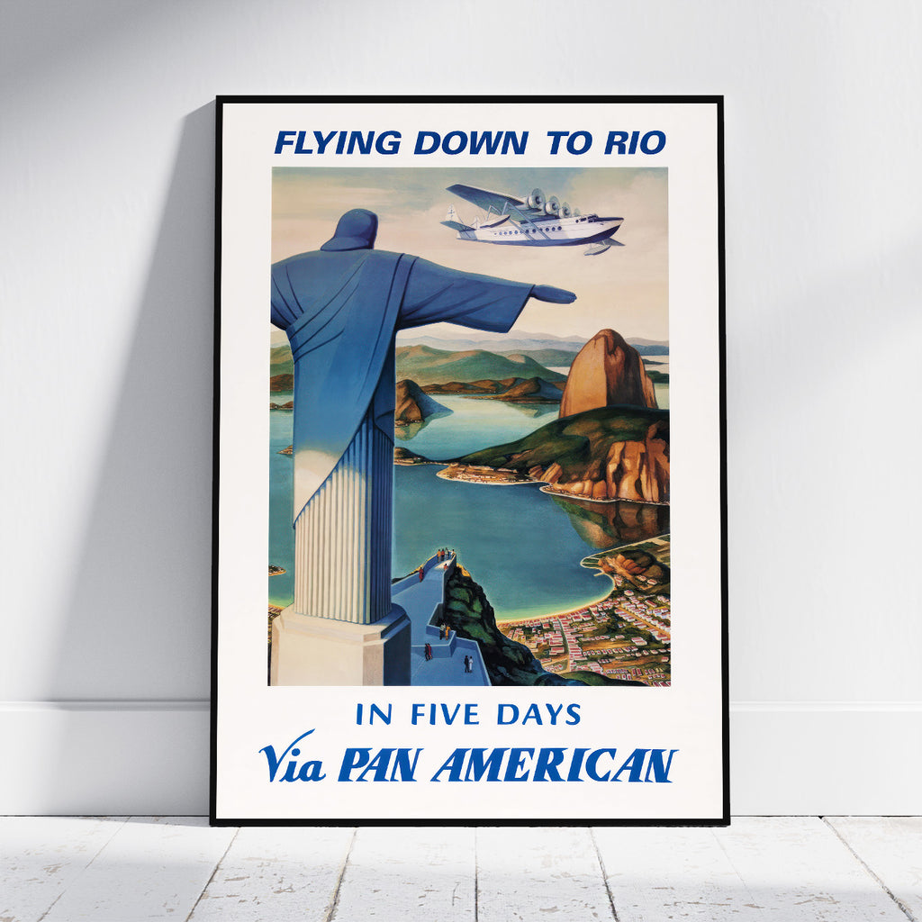 A framed vintage-style travel poster leaning against a white wall on a light floor. The poster reads 'FLYING DOWN TO RIO' at the top and 'IN FIVE DAYS Via PAN AMERICAN' at the bottom, with an illustration of the Rio de Janeiro Christ the Redeemer statue, a seaplane, and coastal scenery.