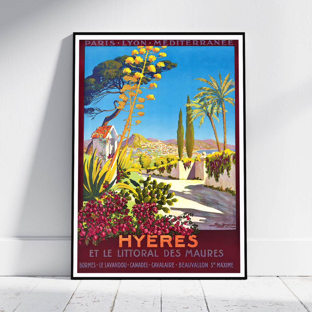 A framed vintage-style travel poster leaning against a white wall on a light floor. The artwork depicts a sunny coastal scene labeled 'HYÈRES ET LE LITTORAL DES MAURES' with blue sky, palm trees, flowers, and a pathway.