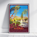 A framed vintage-style travel poster leaning against a white wall on a light floor. The artwork depicts a sunny coastal scene labeled 'HYÈRES ET LE LITTORAL DES MAURES' with blue sky, palm trees, flowers, and a pathway.