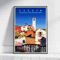 A framed travel poster of Tessin, Switzerland, leaning against a white wall on a light wooden floor. The poster features a colorful illustration of the town's buildings, a prominent white bell tower, and a bright blue sky, enclosed in a thin black frame.