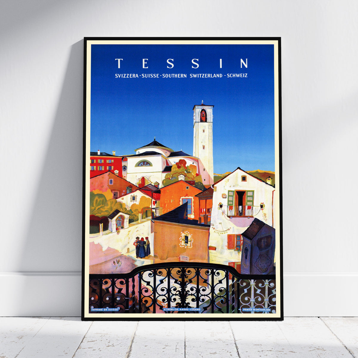 A framed travel poster of Tessin, Switzerland, leaning against a white wall on a light wooden floor. The poster features a colorful illustration of the town's buildings, a prominent white bell tower, and a bright blue sky, enclosed in a thin black frame.