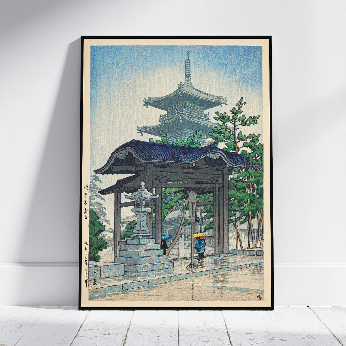 A framed art print of a Japanese ukiyo-e woodblock print depicting a pagoda, gate, stone lantern, and trees, leaning against a white wall on a light wood floor.