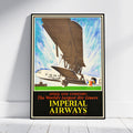 A framed vintage travel poster for Imperial Airways, leaning against a white wall on a light wood floor. The poster features an illustration of an early airplane and a large dirigible balloon, with bold text reading 'IMPERIAL AIRWAYS' and 'SPEED AND COMFORT'.