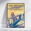 A framed vintage-style travel poster for Swizzair leaning against a white wall on a light wood floor. The poster features a yellow background, a blue airplane, passengers boarding via an airstair, and stylized text including 'čSR-śVżARY', 'ZA 2 HODINY', 'TRATĜ PRAHA-CURYCH', and 'Swizzair'.