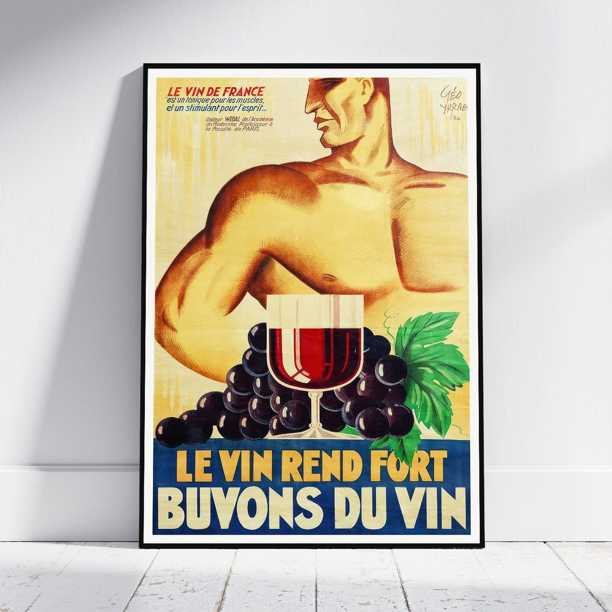 A framed vintage-style French wine poster leaning against a white wall on a light wood floor. The poster shows a shirtless muscular man, a glass of red wine, dark grapes, green leaves, and bold text reading 'LE VIN REND FORT BUVONS DU VIN'.
