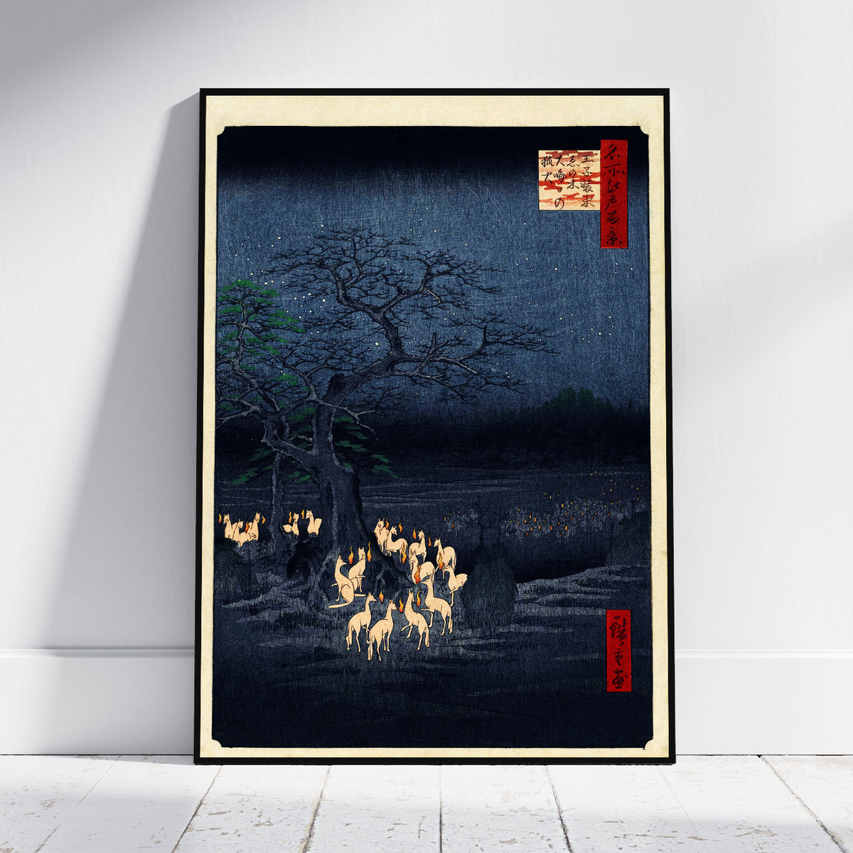 A framed Japanese-style art print leaning against a white wall on a light floor. The artwork depicts a dark night landscape with a large tree, glowing deer, and small red label details, enclosed in a black frame with a cream border.