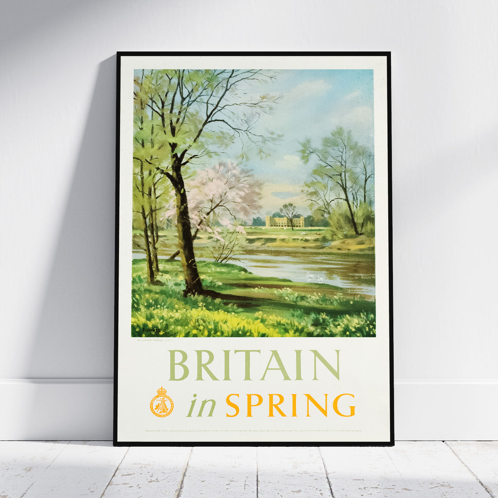 A framed vintage-style travel poster leaning against a white wall on a light wood floor. The poster shows a spring landscape with trees, a river, and a building, with the text 'BRITAIN in SPRING' at the bottom.