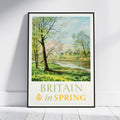 A framed vintage-style travel poster leaning against a white wall on a light wood floor. The poster shows a spring landscape with trees, a river, and a building, with the text 'BRITAIN in SPRING' at the bottom.