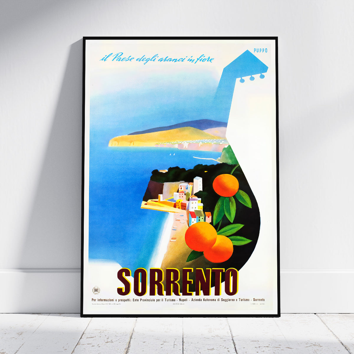 A framed vintage-style travel poster for Sorrento, Italy, leaning against a white wall on a light wood floor. The poster features a stylized guitar silhouette filled with a coastal landscape illustration, including blue sea, yellow cliffs, white buildings, green leaves, and orange fruits, with the word 'SORRENTO' in large letters at the bottom.