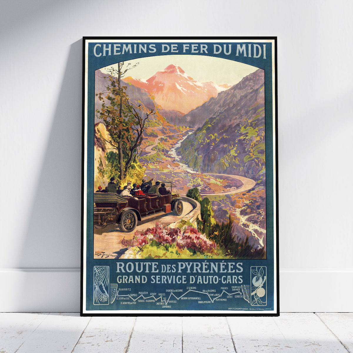 A framed vintage-style travel poster leaning against a white wall on a light wood floor. The poster depicts a mountain valley scene with a car on a winding road and French text reading 'CHEMINS DE FER DU MIDI' and 'ROUTE DES PYRÉNÉENS GRAND SERVICE D'AUTO-CARS'.
