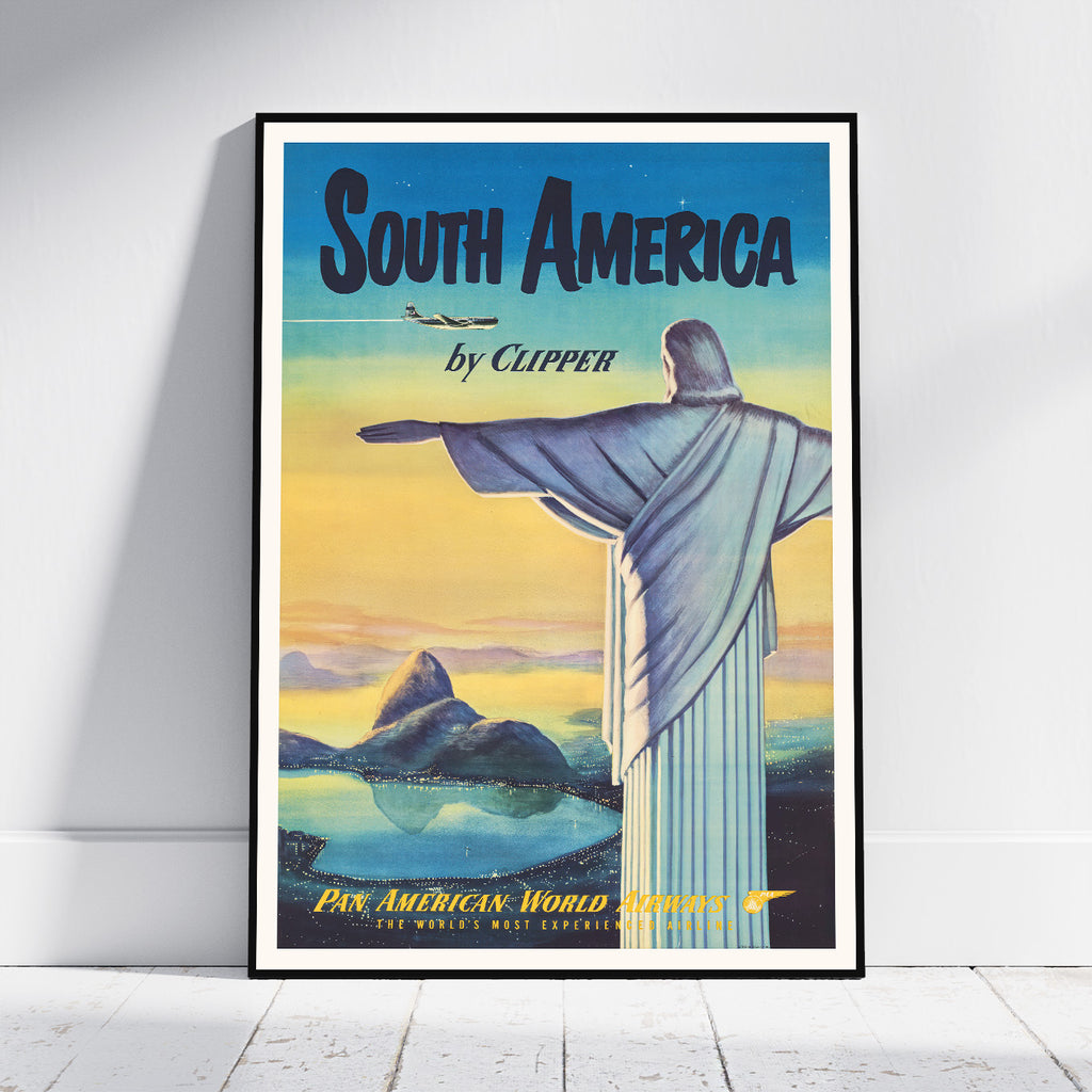 A framed vintage-style travel poster for South America leaning against a white wall on a light wood floor. The poster features the Christ the Redeemer statue, a mountain, a plane, and the text 'South America by Clipper' with 'Pan American World Airways' at the bottom.