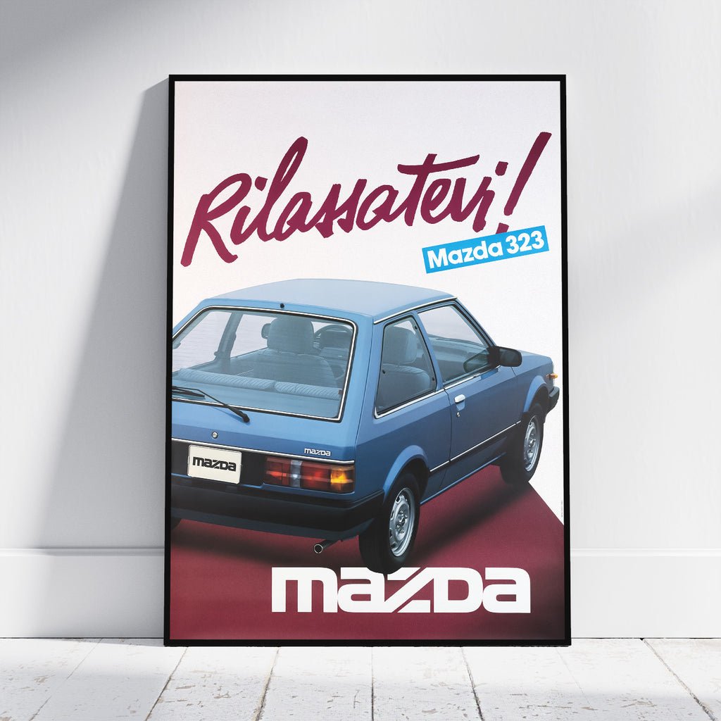 A framed vintage-style poster leaning against a white wall on a light floor. The poster features a blue Mazda 323 car and large Italian text reading 'Rilassatevi!' with 'Mazda' at the bottom.