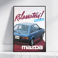 A framed vintage-style poster leaning against a white wall on a light floor. The poster features a blue Mazda 323 car and large Italian text reading 'Rilassatevi!' with 'Mazda' at the bottom.