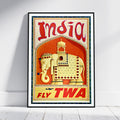 A framed vintage-style travel poster for India, leaning against a white wall on a light floor. The poster features a cream-colored elephant with red and gold decorative details on a warm orange-red background, with large red 'India' lettering at the top and 'FLY TWA' at the bottom.