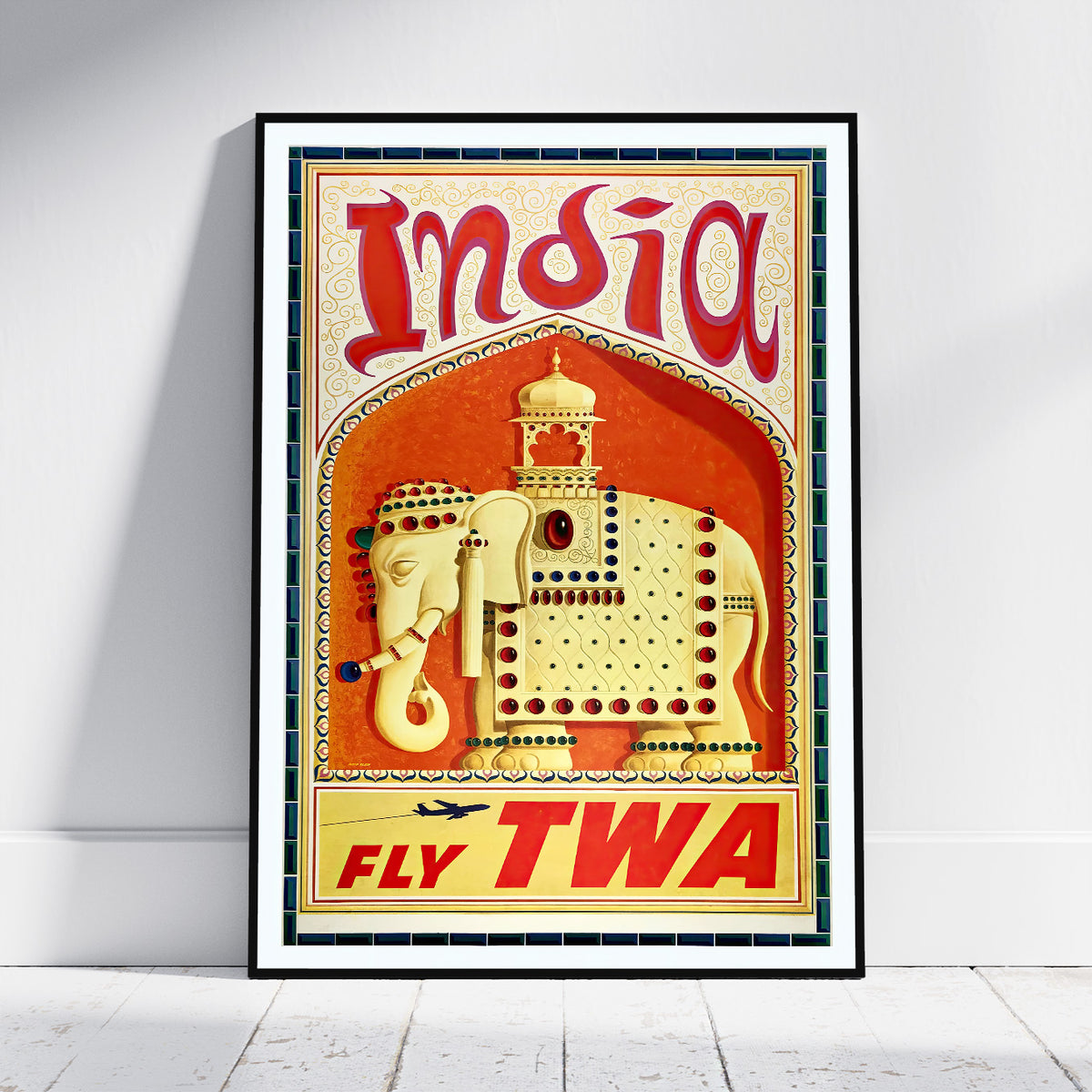 A framed vintage-style travel poster for India, leaning against a white wall on a light floor. The poster features a cream-colored elephant with red and gold decorative details on a warm orange-red background, with large red 'India' lettering at the top and 'FLY TWA' at the bottom.