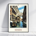 A framed Venice canal print leaning against a white wall on a light floor. The artwork shows gondolas on the water, historic buildings, and the Bridge of Sighs over the canal, with the word 'VENEZIA' printed at the bottom.
