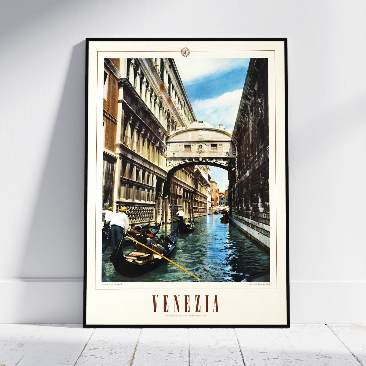 A framed Venice canal print leaning against a white wall on a light floor. The artwork shows gondolas on the water, historic buildings, and the Bridge of Sighs over the canal, with the word 'VENEZIA' printed at the bottom.