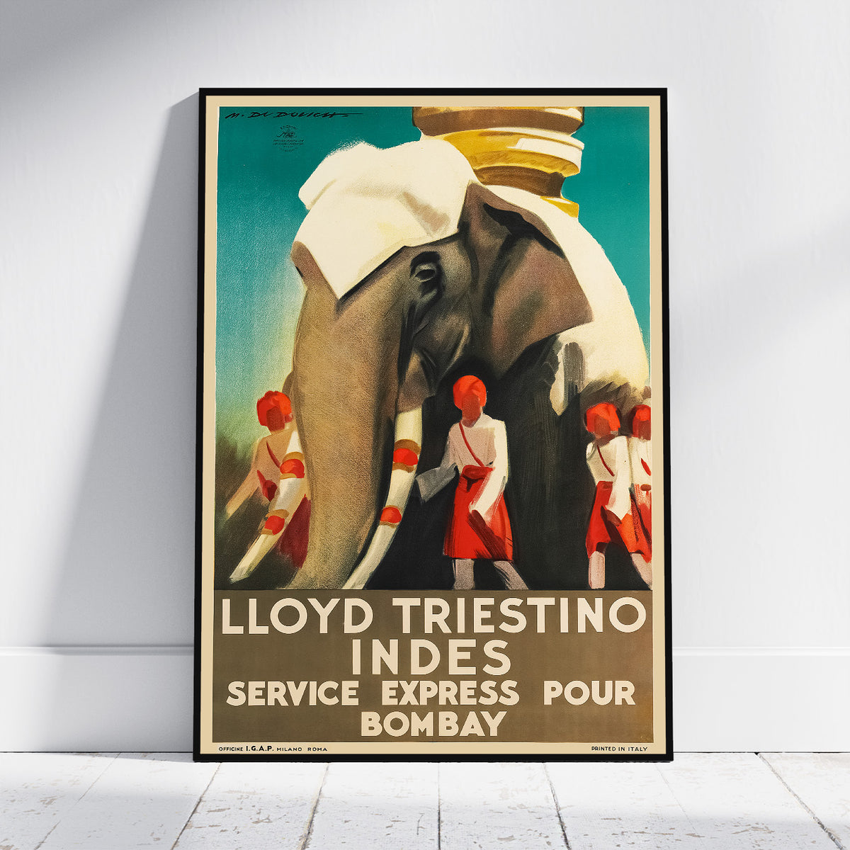 A framed vintage travel poster leaning against a white wall on a light floor. The poster shows an elephant draped in white cloth with several uniformed attendants in red and white, and large text reading 'LLOYD TRIESTINO INDES SERVICE EXPRESS POUR BOMBAY'.