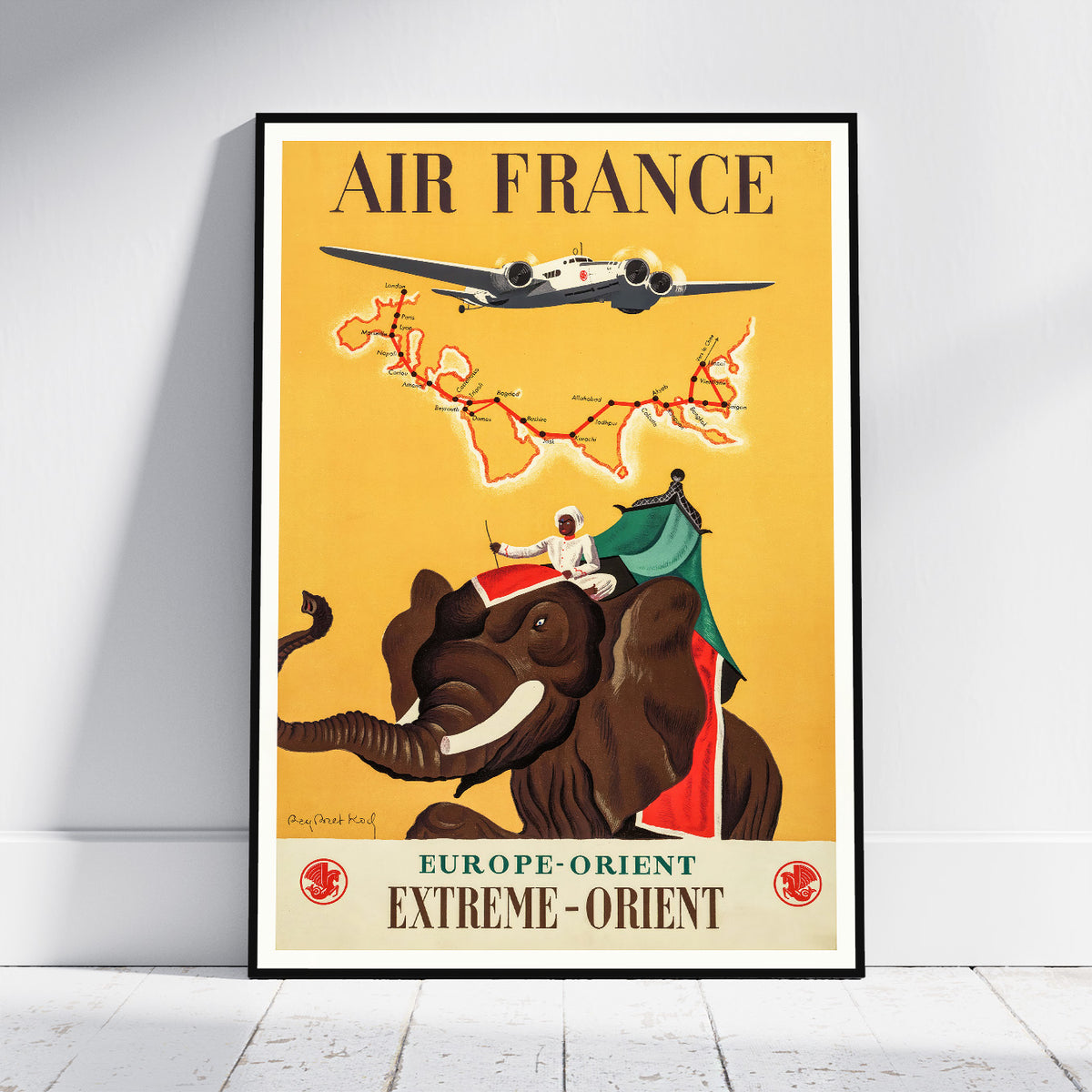 A framed vintage-style Air France travel poster leaning against a white wall on a light floor. The poster features the words 'AIR FRANCE' at the top, an airplane flying above a map, and an elephant carrying a rider with green and red fabric.