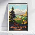 A framed vintage travel poster for the Banff Canadian Pacific Railway, leaning against a white wall on a light wood floor. The poster features a scenic illustration of a mountain valley with a hotel, evergreen trees, and two people, with 'Banff' and 'CANADIAN PACIFIC RAILWAY' printed at the bottom.