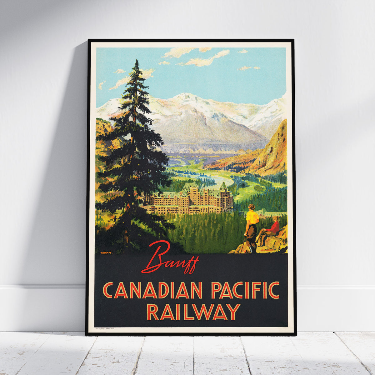 A framed vintage travel poster for the Banff Canadian Pacific Railway, leaning against a white wall on a light wood floor. The poster features a scenic illustration of a mountain valley with a hotel, evergreen trees, and two people, with 'Banff' and 'CANADIAN PACIFIC RAILWAY' printed at the bottom.