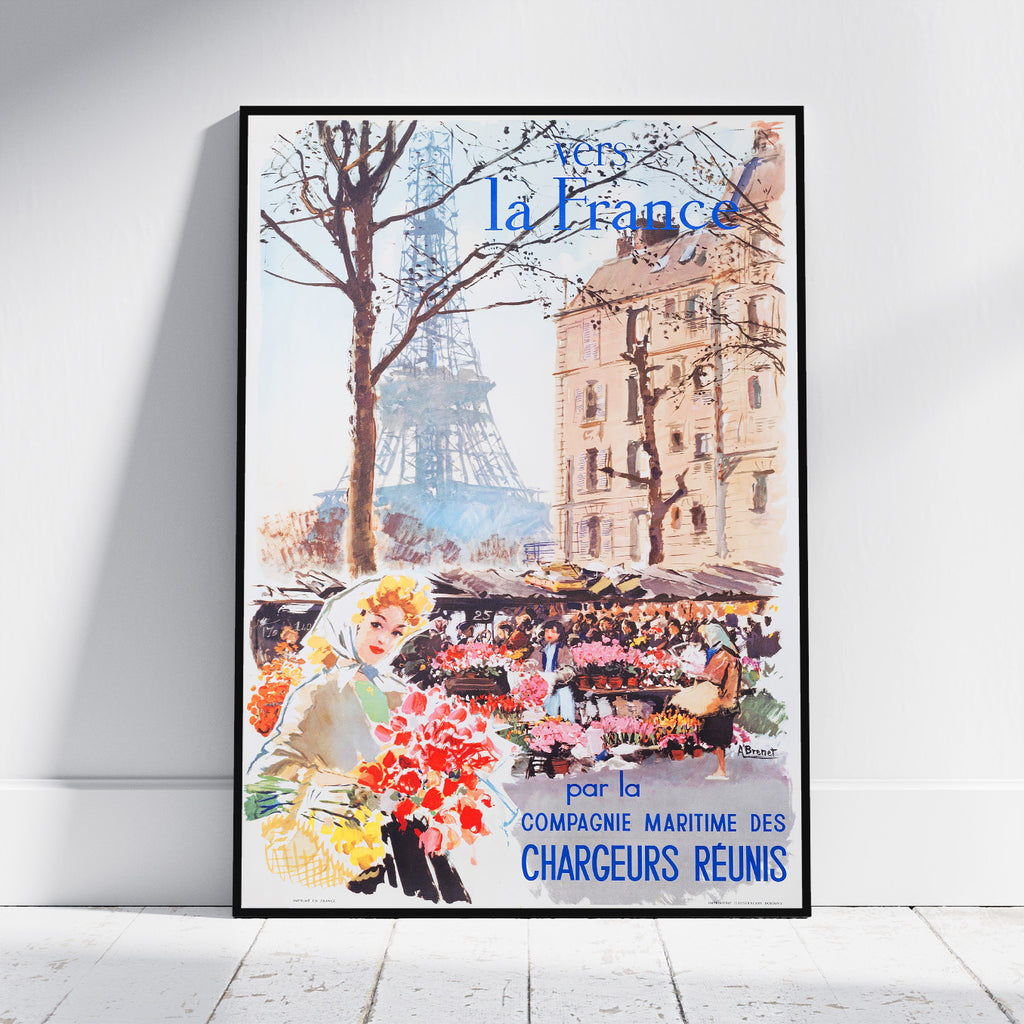 A framed vintage-style travel poster leaning against a white wall on a light floor. The poster depicts a Paris street scene with the Eiffel Tower, trees, a building, flower markets, and a woman holding a bouquet.