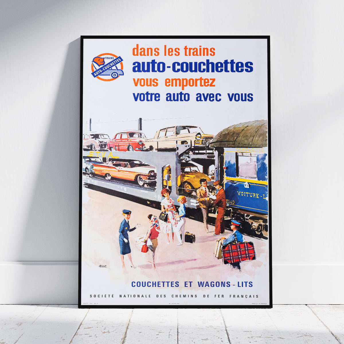 A framed vintage French travel poster leaning against a white wall on a light floor. The poster features retro illustration of trains, cars, passengers, and French text reading 'dans les trains auto-couchettes vous emportez votre auto avec vous' and 'COUCHETTES WAGONS-LITS'.