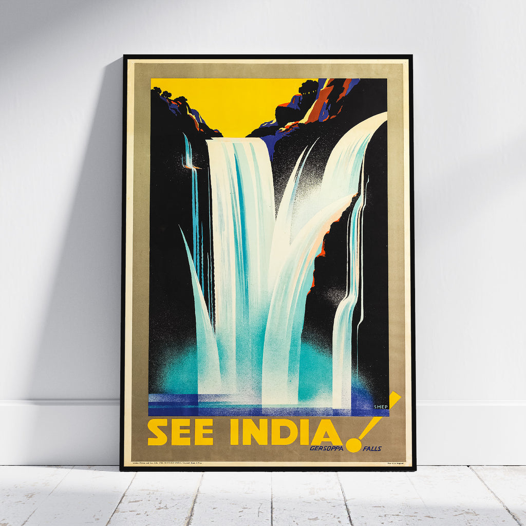 A framed vintage-style travel poster leaning against a white wall on a light floor. The poster depicts a waterfall scene with bold yellow, black, blue, and white colors and the text 'SEE INDIA!' at the bottom.