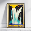 A framed vintage-style travel poster leaning against a white wall on a light floor. The poster depicts a waterfall scene with bold yellow, black, blue, and white colors and the text 'SEE INDIA!' at the bottom.