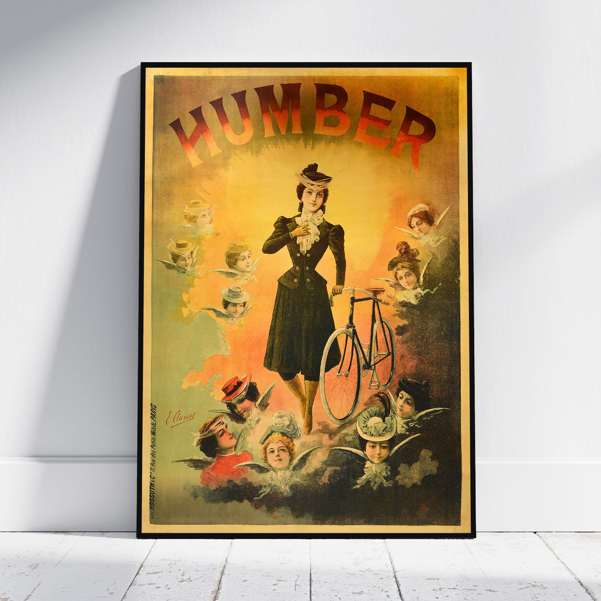 A framed vintage-style poster leaning against a white wall on a light floor. The poster features the word “HUMBER” in large red letters at the top and an illustrated woman in a dark dress standing beside a bicycle, surrounded by smaller figures and objects in warm yellow, orange, green, and dark blue tones.