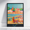 A framed art print featuring a colorful, stylized illustration of a beach and harbor scene with people, buildings, domes, and boats. The print is displayed in a black frame against a white wall on a light wood floor.