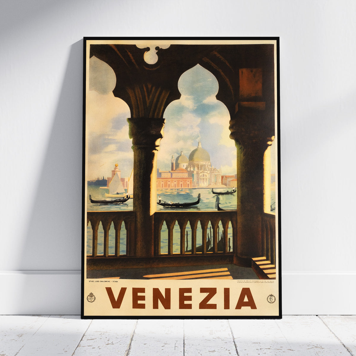 A framed vintage-style travel poster of Venice, leaning against a white wall on a light floor. The artwork shows an arched balcony overlooking gondolas and the Venetian skyline, with the word “VENEZIA” in large brown letters at the bottom.