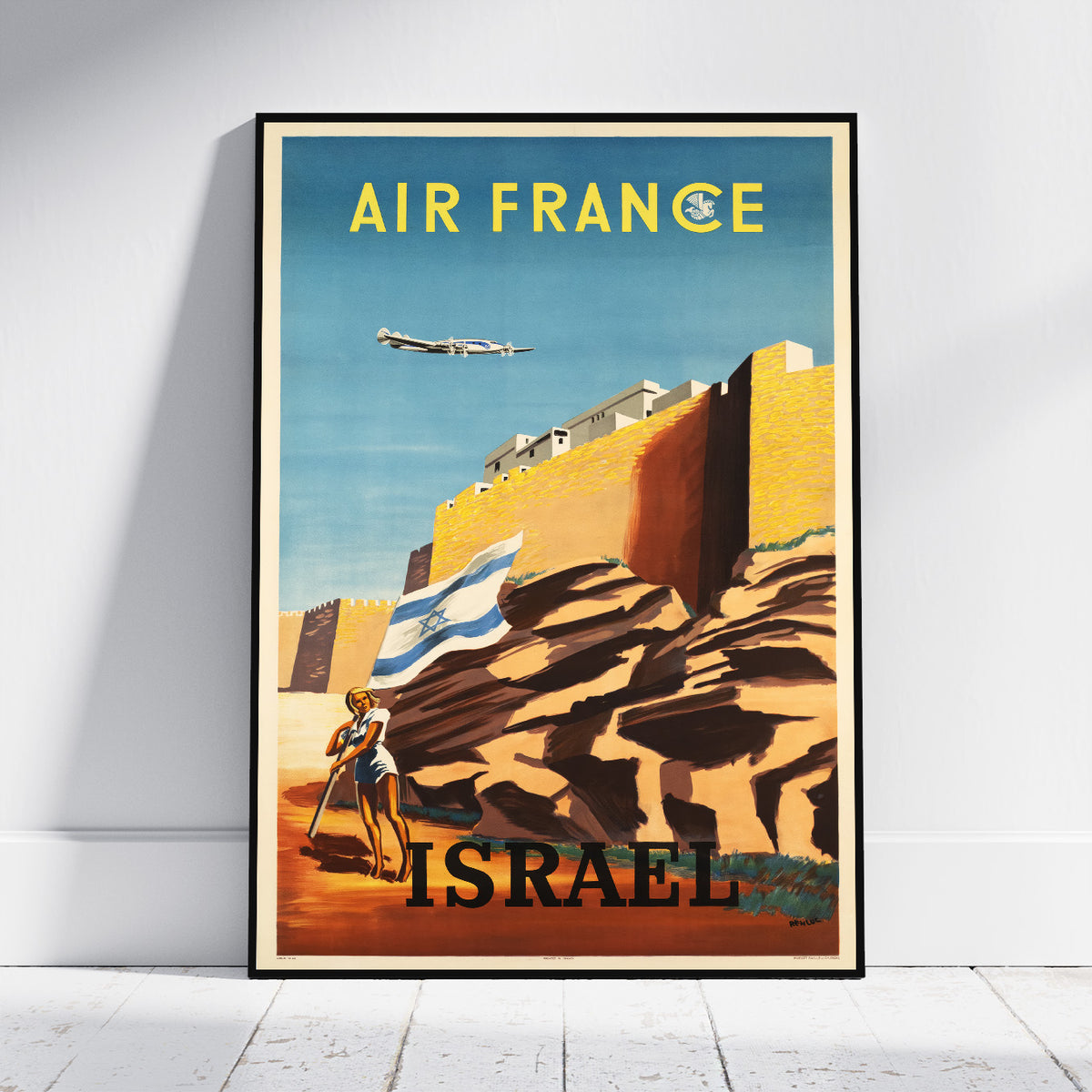 A framed vintage-style travel poster leaning against a white wall on a light floor. The poster reads 'AIR FRANCE' at the top and 'ISRAEL' at the bottom, showing a blue sky, yellow-orange rock formations, an airplane, and a person carrying an Israeli flag.