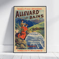 A framed vintage travel poster for Allevard Les Bains (Isere) is shown leaning against a white wall on a light wood floor. The poster features a colorful illustration of people in a red dress and a brown jacket against a mountain and thermal spa background.