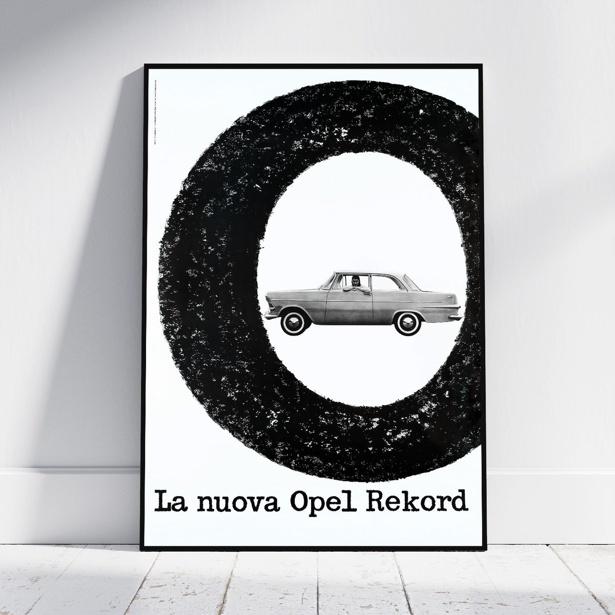 A framed black and white poster leaning against a white wall on a light floor. The poster features a large textured black ring with a vintage car centered inside, and the text 'La nuova Opel Rekord' at the bottom.