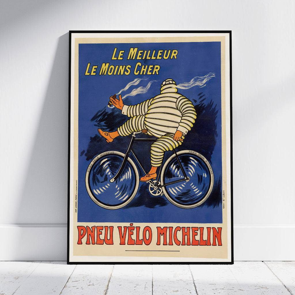 A framed vintage-style Michelin poster leaning against a white wall on a light floor. The artwork shows the Michelin Man riding a bicycle with French text reading 'Le Meilleur Le Moins Cher' and 'Pneu Vélo Michelin'.
