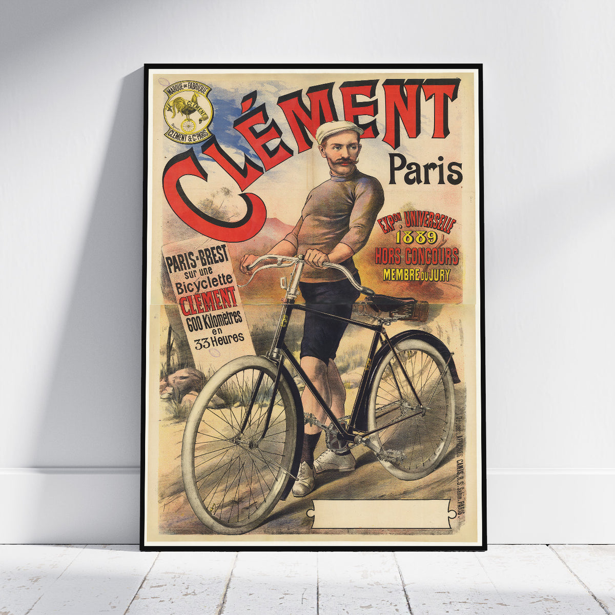 A framed vintage-style poster leaning against a white wall on a light wood floor. The poster features the text 'Clement Paris' and an illustration of a man riding a bicycle, with retro typography and warm cream, red, black, and tan tones.
