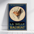 A vintage-style framed poster leaning against a white wall on a light floor. The poster features an illustrated sleeping figure inside a cream oval on a dark blue background, with French text reading 'PLUS DOUCE QU'UN OREILLER !' at the top and 'LA SELLE BAURIAT' in large yellow letters at the bottom.