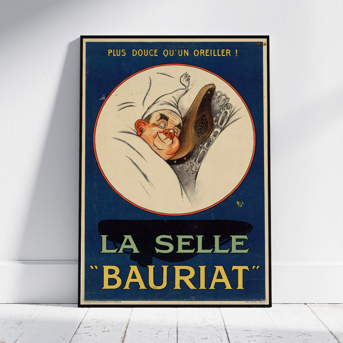 A vintage-style framed poster leaning against a white wall on a light floor. The poster features an illustrated sleeping figure inside a cream oval on a dark blue background, with French text reading 'PLUS DOUCE QU'UN OREILLER !' at the top and 'LA SELLE BAURIAT' in large yellow letters at the bottom.
