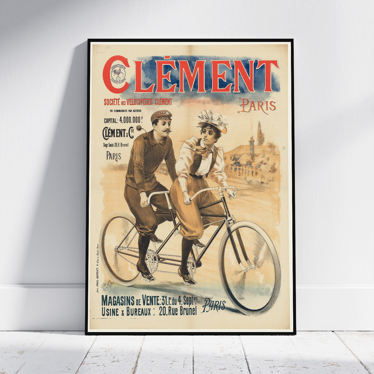 A framed vintage-style poster leaning against a white wall on a light wood floor. The poster features the text “Cément Paris” at the top and an illustration of two people riding a tandem bicycle, with additional aged-paper typography and scenery in the background.