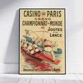 A framed vintage sports poster leaning against a white wall on a light wood floor. The poster reads 'CASINO DE PARIS GRAND CHAMPIONNAT DU MONDE DES JOUTES A LA LANCE' and shows illustrated runners in boats with a red racing boat and cream background.