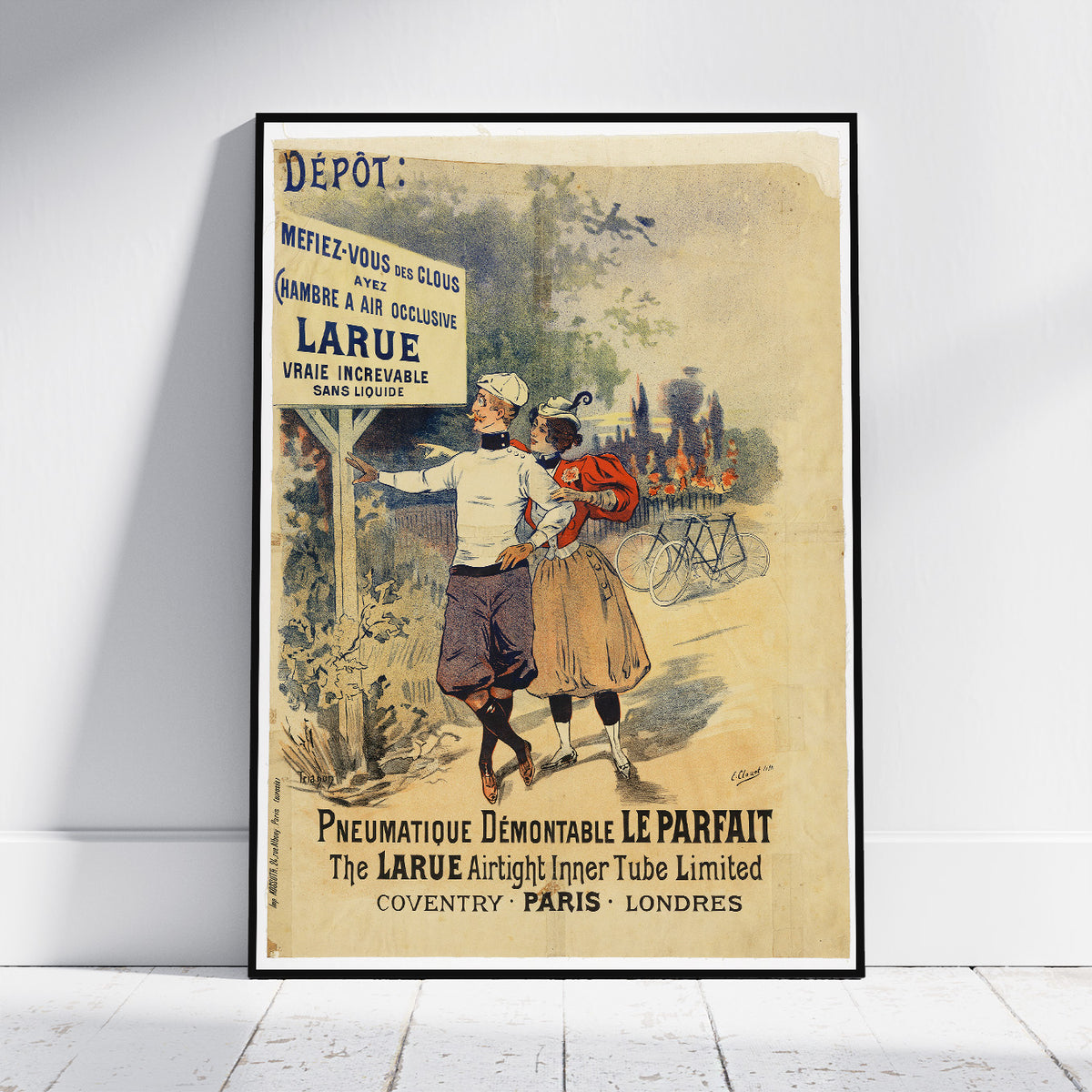 A vintage-style framed poster leaning against a white wall on a light wood floor. The poster features an illustrated scene with a man and a child, bicycles, and large text including 'LARUE' and 'Pneumatique Démontable Le Parfait.