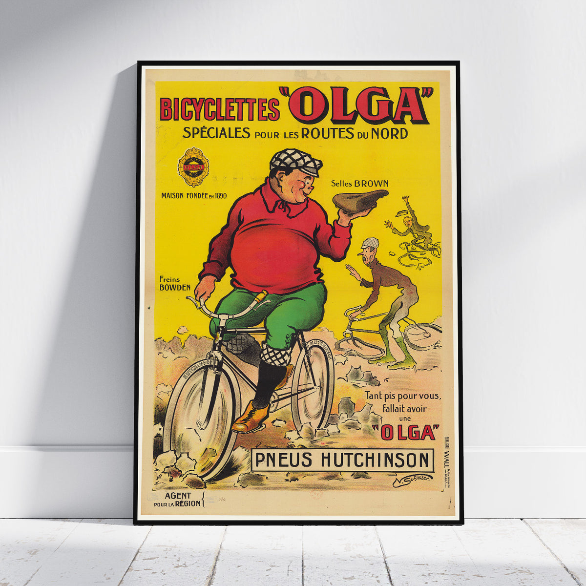 A framed vintage-style bicycle poster leaning against a white wall on a light wood floor. The poster features a yellow background with illustrated text and a cyclist in a red top and green pants, plus visible text including 'BICYClettes "OLGA"' and 'Pneu Hutchinson'.