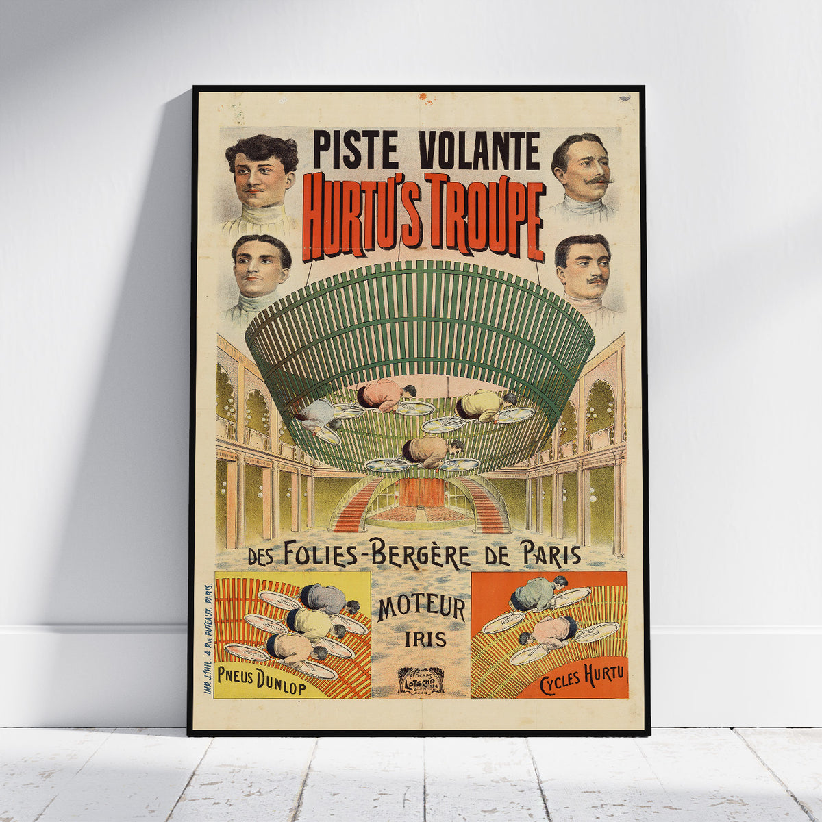 A framed vintage poster for 'Piste Volante Hurty's Troupe' leaning against a white wall on a light wooden floor. The poster features a large green flying machine illustration with portraits of men and French text advertising Parisian flying shows.